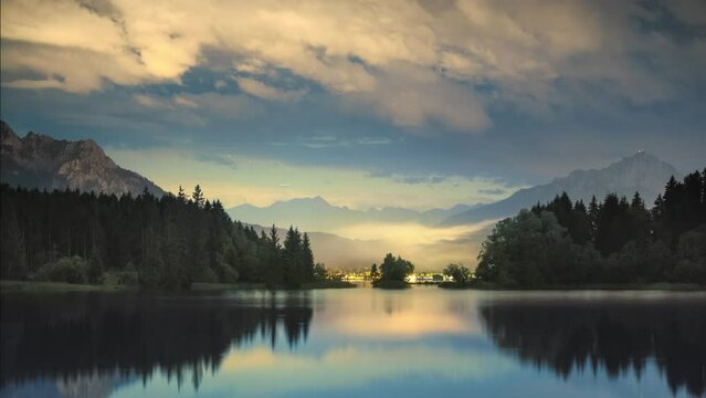 Time lapse of illuminated clouds over a scenic lake at night. Picturesque mountain landscape with blue atmosphere and tree silhouettes reflected in the water

