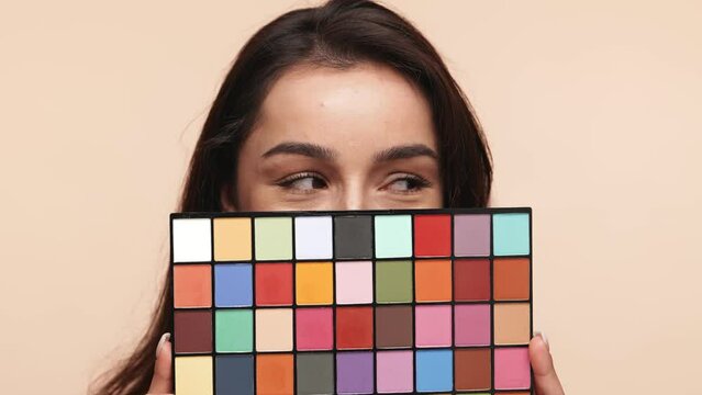 Close-up of a young woman with striking eyes peeking over a vibrant makeup palette, conveying options and personalization in beauty on a beige background.