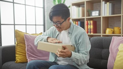 A smiling asian man unboxing a package while sitting in a modern living room, expressing joy and curiosity.