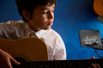 Hispanic teenage boy playing acoustic guitar at home, sitting against blue wall with musical...