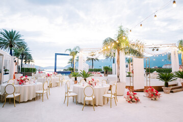 Covered festive tables with bouquets of flowers and garlands of light bulbs stand on the terrace