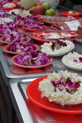 Lined up white and purple blossoms and different fruits on red dishes and silver tablets as offerings at a temple in Asia.