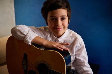 Teenager boy, a little guitarist musician holding acoustic guitar, smiling looking at camera,...