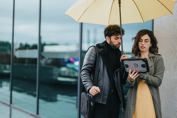 Two modern business executives examining a digital tablet under a shared yellow umbrella, with an...