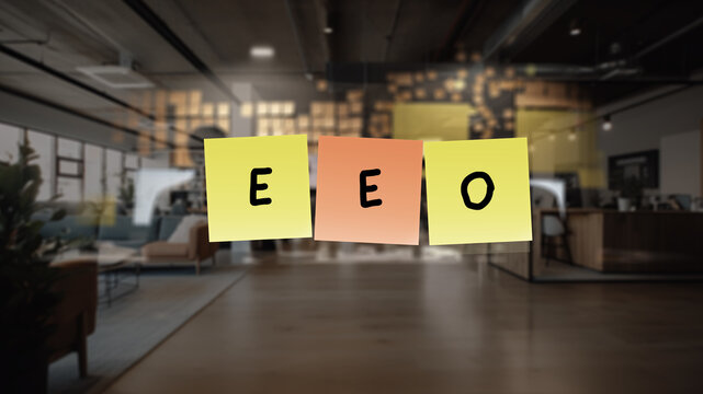EEO written in black marker. The notes are affixed to a transparent surface in front of open office, three sticky notes, each with a single letter .
