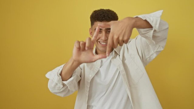 Cheerful young arab man framing a photo with hands, radiating creativity on a vibrant yellow isolated background