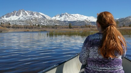 Fototapeta premium A woman with long, red hair sits in a boat on a lake against a mountain range in the background