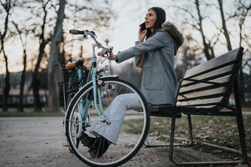 Elegant businesswoman with bicycle taking a phone call on a park bench during the evening.