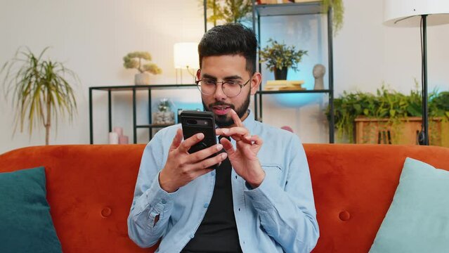 Surprised excited Arabian young man winner holding smartphone reading good news amazed by online bet bid game win. Happy Indian guy on sofa looking at screen overjoyed by victory success at home.