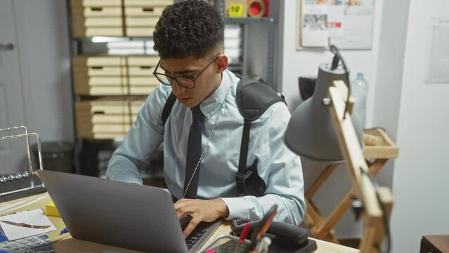 Young man concentrates on a laptop in an office with evidence, evoking a detective's workspace.