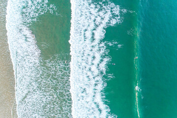 Foam waves breaking near the beach, turquoise sea and two surfers, seen from a drone. Aerial...