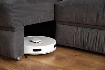 A white robot hoover cleans wood laminate on the floor. collects dust near the sofa in difficult places and leaves from under the large sofa. infrared sensors and cameras control. parktronic.