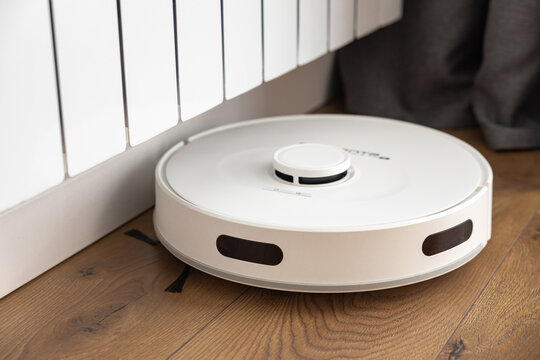 A white robot vacuum cleaner with a sweeping brush, infrared motion sensors and a camera cleans the floor near a heating radiator in a room. domestic helper