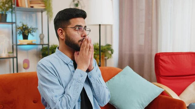 Tensed Hispanic man sits on sofa at home folding palms together pray ask for help thinks feel stressed looks desperate by life problems. Worried upward Indian guy praying making wish begging apology