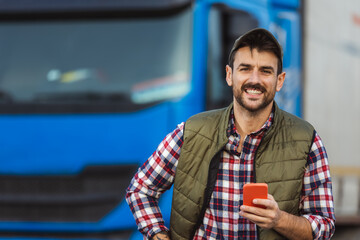 Happy confident male driver standing in front on his truck, using his phone.