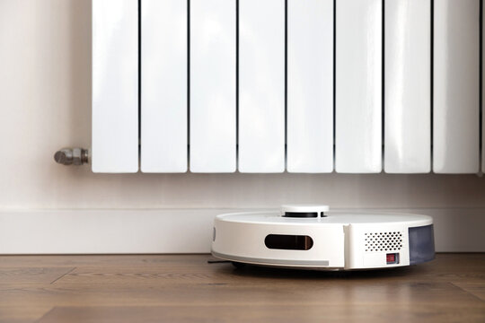 A white robot vacuum cleaner with a sweeping brush, infrared motion sensors and a camera cleans the floor near a heating radiator in a room. domestic helper