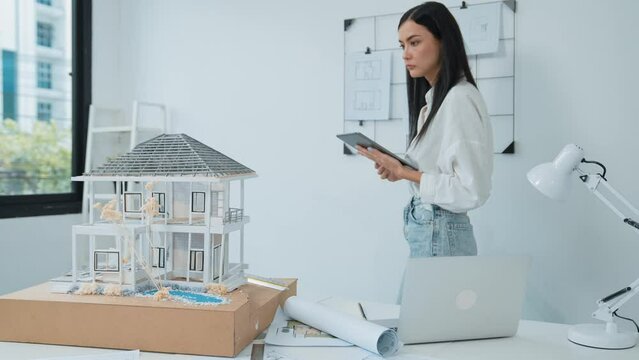 Beautiful caucasian architect focus on using tablet to check house model on meeting table with blueprint, house model and architectural document scatter around at meeting room. Immaculate.