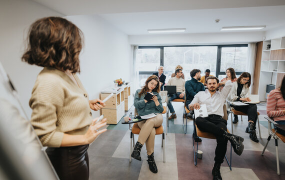Diverse group of professionals in a business workshop. A speaker leads the casual meeting while attendees are seated, some taking notes and one man smiling at the camera.
