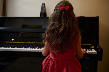 Back view of a little kid girl in elegant red dress, sitting at piano forte, performing classical...