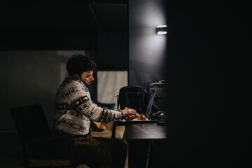 A solitary man concentrated on work using his laptop late at night in a dimly lit environment,...