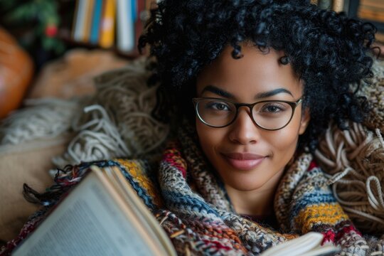 Beautiful young woman reading a book while sitting on a sofa in a cozy room. Charming African American girl has covered herself with a warm blanket and spends a long winter evening reading.