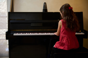 View from the back of little child girl in stylish red dress, playing grand piano at home. Talented...