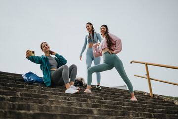 Three fitness enthusiasts enjoy a break to take a selfie on stairs, showcasing workout friendship and fun.
