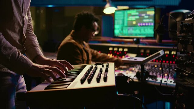 Musician playing piano midi controller in control room at professional recording studio, creating sounds with electronic keyboard notes. Sound engineer does mix and master on files. Camera B.