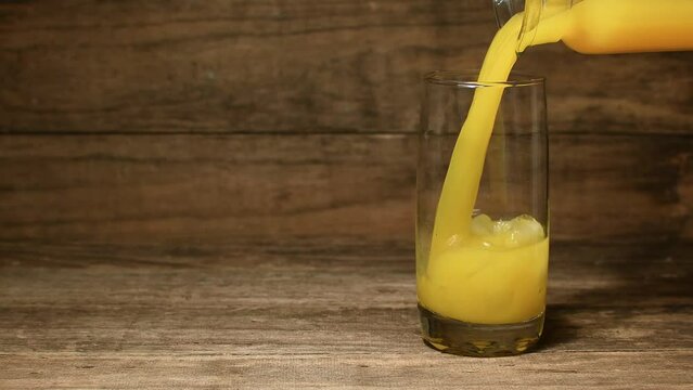 Medium, low shot, glass jug pouring orange juice into a glass with ice, brown, rustic wooden setting, copy space to the left