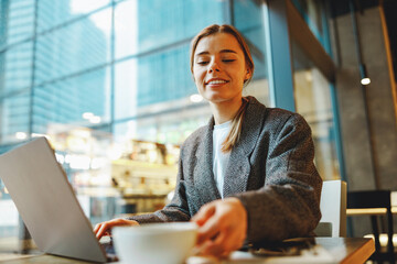 Smiling business woman is drinking coffee in coworking while working on laptop near window