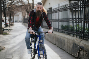 A bearded businessman in casual attire rides his bicycle in an urban setting, reflecting a modern, eco-conscious approach to city commuting and remote working.