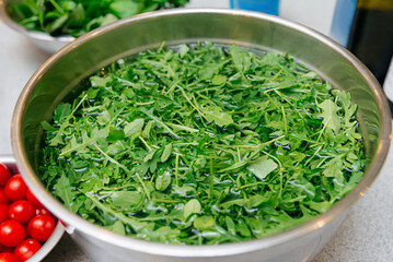 Fresh Arugula Leaves in Stainless Steel Bowl. A large bowl filled with vibrant green arugula leaves soaking in water, prepared for cooking or salad making, on a kitchen countertop.