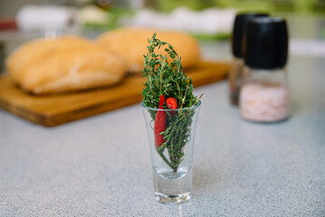 Fresh Herbs in Glass and Baked Bread on Kitchen Counter. A transparent glass filled with fresh thyme and a red chili pepper, beside freshly baked loaves of bread and kitchen seasoning essentials.