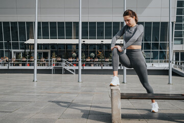 Focused young woman in sportswear performs leg stretches on a sunny day in a modern urban setting, exemplifying healthy lifestyle choices.