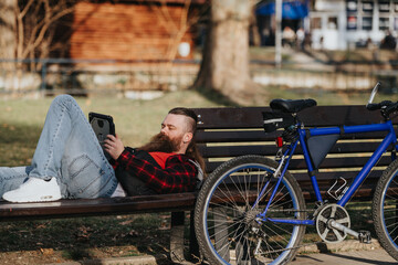 A bearded businessman in casual attire enjoys remote work in an urban park setting, using a tablet...