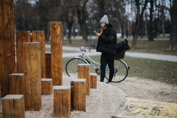 Woman using smart phone near bike in park with wooden playground installation.