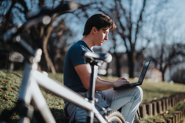 A focused man sits on a bench with a bicycle next to him, engaged in work on his laptop in a park.
