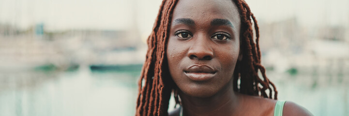 Panorama of woman with African braids wearing top looks at camera on the yachts and ships background, standing on the pier in the port.
