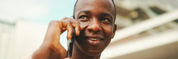 Smiling young african student sitting outside of university talking on cellphone, Close-up, Panorama.
