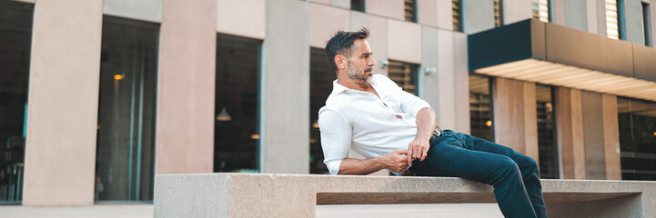 Mature businessman with neat beard wearing white shirt sits on bench on the street of modern city,...