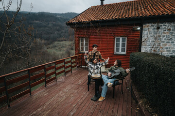 Three friends share a moment of music and relaxation on the porch of a cozy wooden cabin surrounded by nature.