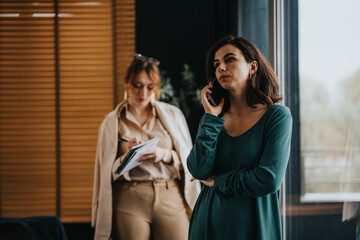 Two professional women engaged in a project meeting, with one taking notes and the other on a phone call discussing important details, costs, and solutions.
