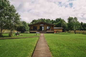 Fototapeta na wymiar Wooden Cabin with Pathway in Green Landscape. A wooden cabin retreat is nestled in a lush green landscape, complete with a welcoming boardwalk and surrounding trees.