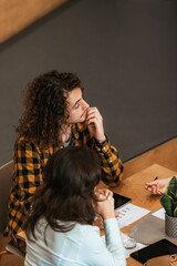 Engaged curly-haired woman in a plaid shirt participating in a business meeting with thoughtful expression, discussing charts and strategies with coworker.