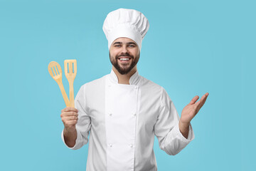 Happy young chef in uniform holding wooden utensils on light blue background