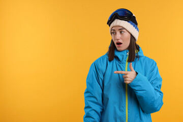 Winter sports. Surprised woman with snowboard goggles pointing at something on orange background,...