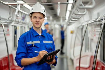 A confident railway technician wearing a safety helmet is holding a digital tablet, standing inside...