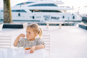 Little girl eats ice cream with a spoon, holding a glass with her hand on a table on the seashore