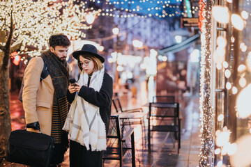 Young couple using smart phone on festive city street decorated with Christmas lights.