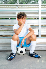 Bored young soccer player sitting on a soccer ball waiting for his chance to play the game